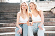 Two young beautiful smiling hipster female in trendy summer white top and jeans clothes. Carefree women posing in the street. Positive models having fun outdoors. Cheerful and happy. Sit at stairs