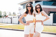 Two young beautiful smiling female in trendy summer white cycling shorts, top clothes. Sexy carefree women posing in street at sunny day. Positive models having fun. Cheerful and happy. In sunglasses