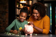Smiling african american mother helping daughter putting money in piggy bank. Cute little black girl saving money by adding a coin in piggy bank with mother at home.