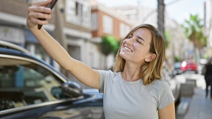 Poster - Joyful young blonde woman confidently making a fun selfie with her smartphone on the city street, basking in sunlight while enjoying her urban lifestyle.