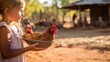 A child holds chicken food in her outstretched palms, towards a tame chicken, who approaches her. Focus is on the red hen. Various other chickens stand in the background.
