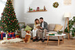 happy couple sitting on couch and reading book near corgi dog and Christmas tree with presents