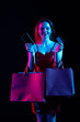 Attractive woman with shopping bags holding smartphone and credit card, looking at camera smiling. Isolated on black background in neon light.