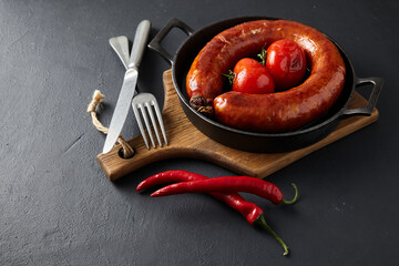 Wall Mural - Ring of fried fatty pork sausage and grilled tomatoes on a black cast iron frying pan on a wooden board on a black stone table