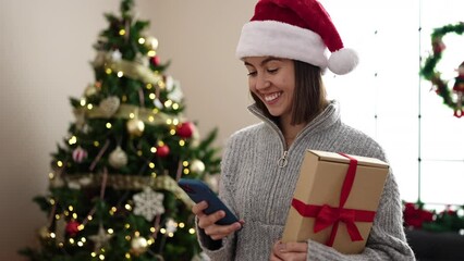 Poster - Young beautiful hispanic woman using smartphone standing by christmas tree at home