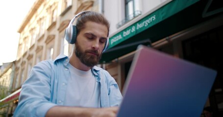 Portrait of young stylish man in headphones sitting in cafe shop at urban city street outdoors. Bearded guy working on computer, sending messages, makes online purchases, watching movies.