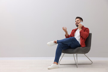 Poster - Handsome man talking on smartphone while sitting in armchair near light grey wall indoors, space for text