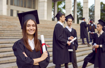 Portrait of young smiling joyful girl student in university graduate gown and diploma in hands. Happy graduated woman standing proudly with crossed arms. Graduation celebration and education concept.