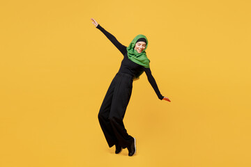 Canvas Print - Full body young arabian asian muslim woman in green hijab abaya black clothes stand on toes with outstretched hand leaning back isolated on plain yellow background People uae islam religious concept