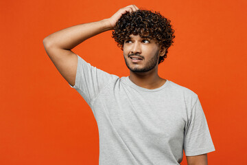 Wall Mural - Young sad mistaken upset puzzled Indian man he wearing t-shirt casual clothes scratch hold head look aside on area mock up isolated on orange red color background studio portrait. Lifestyle concept.