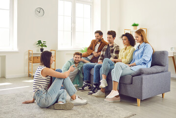 Wall Mural - Group of friends hanging out together. Several young people meeting at somebody's place. Young men and women sitting on sofa and listening to girl who is sitting on floor and talking about something