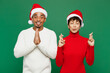 © ViDi Studio - Merry young couple man woman wear red casual clothes Santa hat posing keep fingers crossed hold hand folded in prayer gesture isolated on plain green background. Happy New Year 2024 Christmas concept.