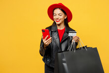 Young Minded Pensive Woman Wears Casual Clothes Red Hat Hold Shopping Paper Package Bags Use Mobile Cell Phone Credit Bank Card Isolated On Plain Yellow Background. Black Friday Sale Buy Day Concept.