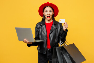 Wall Mural - Young surprised IT woman wear casual clothes red hat hold shopping paper package bags use work on laptop pc computer credit card isolated on plain yellow background. Black Friday sale buy day concept.