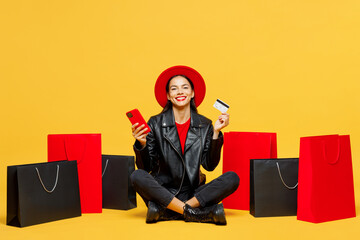 Wall Mural - Full body young woman wear casual clothes red hat sit near shopping paper package bags hold mobile cell phone credit bank card isolated on plain yellow background. Black Friday sale buy day concept.