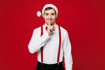 Wall Mural - Merry young fun man wear white shirt Santa hat posing say hush be quiet with finger on lips shhh gesture isolated on plain red background studio. Happy New Year Christmas celebration holiday concept.