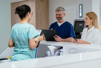 Different age medical team have small meeting at clinics reception. Two doctors talking to receptionist in hospital waiting room. Smiling male doctor writing on document at reception desk.