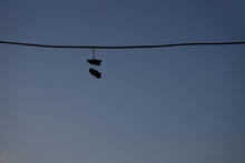 Black Silhouette Of Sneakers, Shoes Hanging From A Telephone Wire On Blue Sky Background. Old Sneakers Hang On An Electric Wire On A Summer Day.