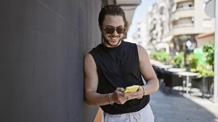 Wall Mural - Young hispanic man wearing sunglasses using smartphone at street