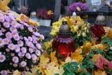 Fototapeta  - Grave with glass candles and flowers in pots among dry autumn leaves in cemetery on November day