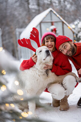 Lovely couple hug with their cute dog wearing toy deers horns at snowy backyard. Young family spending happy winter time together outdoors