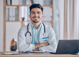 Poster - Happy, portrait and man doctor at desk in a healthcare, medical and hospital office. Smile, male professional and surgeon worker with arms crossed and computer feeling confident and pride in clinic