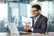 © Liubomir - Young happy and smiling financier inside the office at the workplace, businessman shows joyful to the laptop camera the graph of the achievement of the result, online video meeting with colleagues.