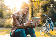 Beautiful Young Smiling Woman Sitting Alone In A City Park And Using Digital Tablet Pc. Blonde Female Wearing Leather Jacket And Cap Reading On Tablet Outdoors.