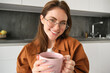 Close up portrait of beautiful woman in glasses, drinks tea, warms her hands with hot cup, smiling and looking happy at camera, sitting in kitchen