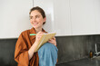 © Mix and Match Studio - Portrait of cute young woman with notebook, sitting in the kitchen and writing her to do list, making tasks for week, puts notes in schedule or planner