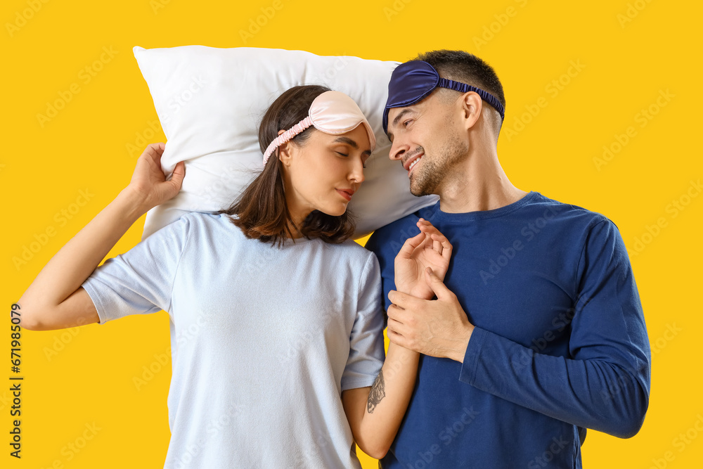 Young couple with sleeping masks and pillow on yellow background