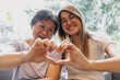 © Suthida - Happy Asian Thai older mother and daughter showing love gesture, smiling making heart sign and looking at camera, sitting on sofa together at home.