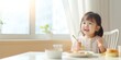 Asian cute little girl holding fork and trying to eat food sitting on the white high chair in the morning at home with copy space. : Generative AI