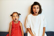 Shocked surprised young female and little girl looking at camera with open mouth and big eyes, astonished with jaw dropping news, sitting on back of couch against white wall at home