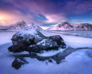 Wall Mural - Sandy arctic beach with blue sea, stone in frozen coast and rocks in snow in winter at sunset in Lofoten islands, Norway. Colorful Landscape with snowy mountains, sky with clouds. Norwegian coast