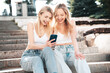 Two young beautiful smiling trendy female. Carefree women posing in street. Positive models having fun. Cheerful and happy. Hold smartphone, look at mobile cellphone screen, use phone apps