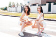 Two young beautiful smiling female in trendy summer white cycling shorts, top clothes. Sexy carefree women posing in street at sunny day. Positive models having fun. Cheerful and happy. Sit at asphalt