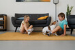 © Studio Marmellata - Side view of focused kids while sitting on carpet with standing pet dog and playing in living room near sofa set potted plant cabinet in daylight at home