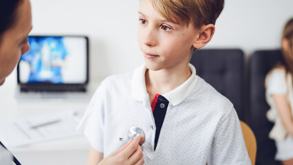 Wall Mural - Doctor and kid patient are in the clinic. Physician in white coat examining a serious young boy with a stethoscope, close up. Medicine, therapy concept