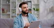 © serg - Attractive man wearing headphones sit on sofa holding tablet device while having a video call on mobile tablet screen talking. E-commerce, virtual services concept