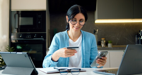 Business woman enters bank credit card digits smartphone to make online purchase. Brunette female online banking with credit card and phone in a home online shopping at home.