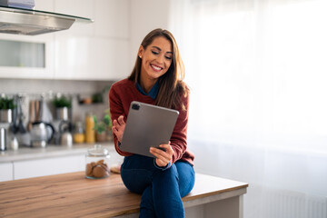 Wall Mural - Smiling woman sitting on kitchen counter at home working on tablet pc. Professional businesswoman looking at tablet screen distantly working from home.