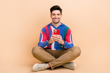 Sticker - Full length photo of smile happy businessman influencer using smartphone surfing social medias sit floor isolated on beige color background