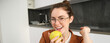 © Mix and Match Studio - Close-up portrait of brunette woman at home, wearing glasses, eating apple in the kitchen and smiling, biting fruit