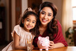 © AI_images - Happy beautiful Indian mother and kid girl saving money, holding piggy bank, looking at camera, smiling, laughing.