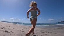Fish Eye Back View To Running Little Girl Along Summer Beach. Wide Shot Of Cute Blonde Girl In Swimsuit Running To Ocean At Sunny Day. Concept Of Let Kids Run On Beach For Healthy Feet And Whole Body