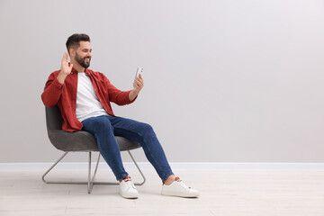 Poster - Handsome man having video chat via smartphone while sitting in armchair near light grey wall indoors, space for text