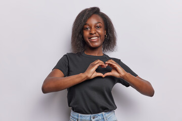 Wall Mural - Horizontal shot of pretty dark skinned woman shows heart sign I love you gesture smiles happily dressed in casual black t shirt and jeans isolated over white background. Romantic feelings concept