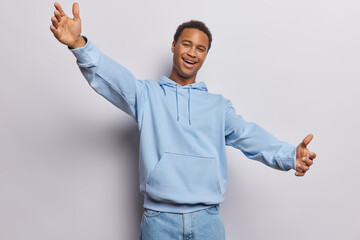 Wall Mural - Positive friendly dark skinned African man keeps arms outstreched wants to hug you dressed in casual blue sweatshirt and jeans meets best friend poses against white background. Come to me closer