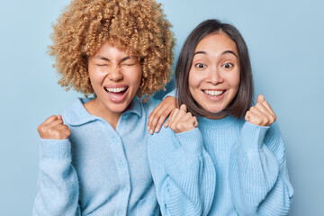 Wall Mural - Horizontal shot of positive young women clench fists and celebrate special occasion dressed in casual jumpers stand closely to each other isolated over blue background. Body language concept